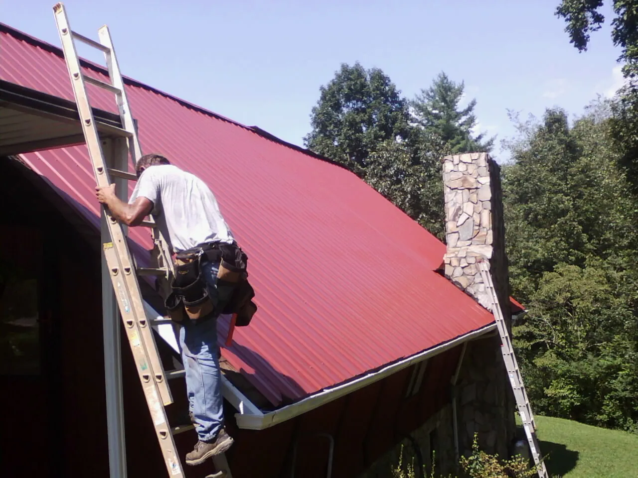Roofer on a ladder beside a red metal roof and stone chimney — Soffit & Fascia Repair by Roofing Brothers in Guilderland Center
